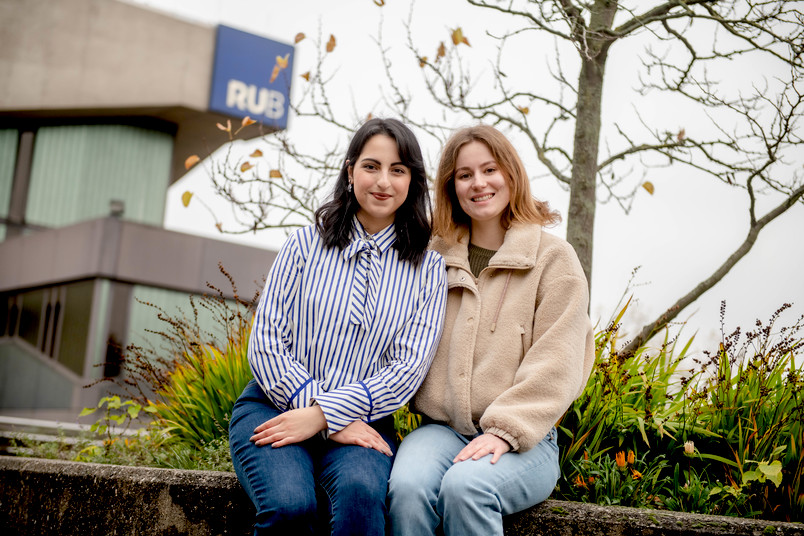 Zwei Studentinnen sitzen nah an einander im grüner Punkt vor dem RUB Logo