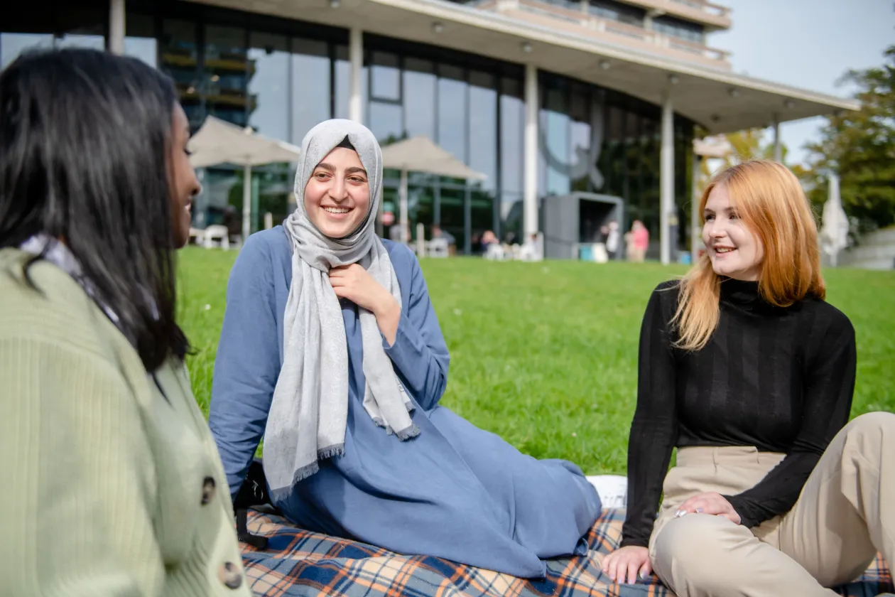 Drei Studierende sitzen auf einer Wiese vor dem QWest und unterhalten sich.