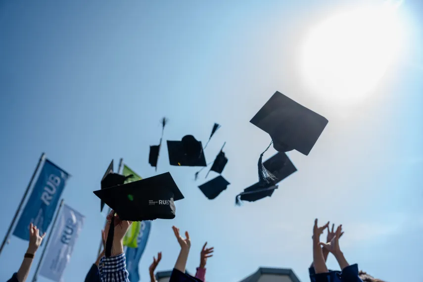 Doktorhüte in die Luft geschmissen, im Hintergrund die RUB-Flagge