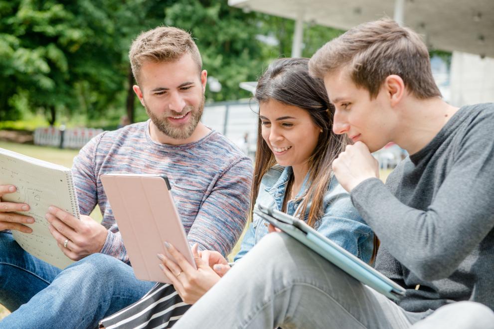 Drei Studierende schauen sich Inhalte auf einem Tablet an.