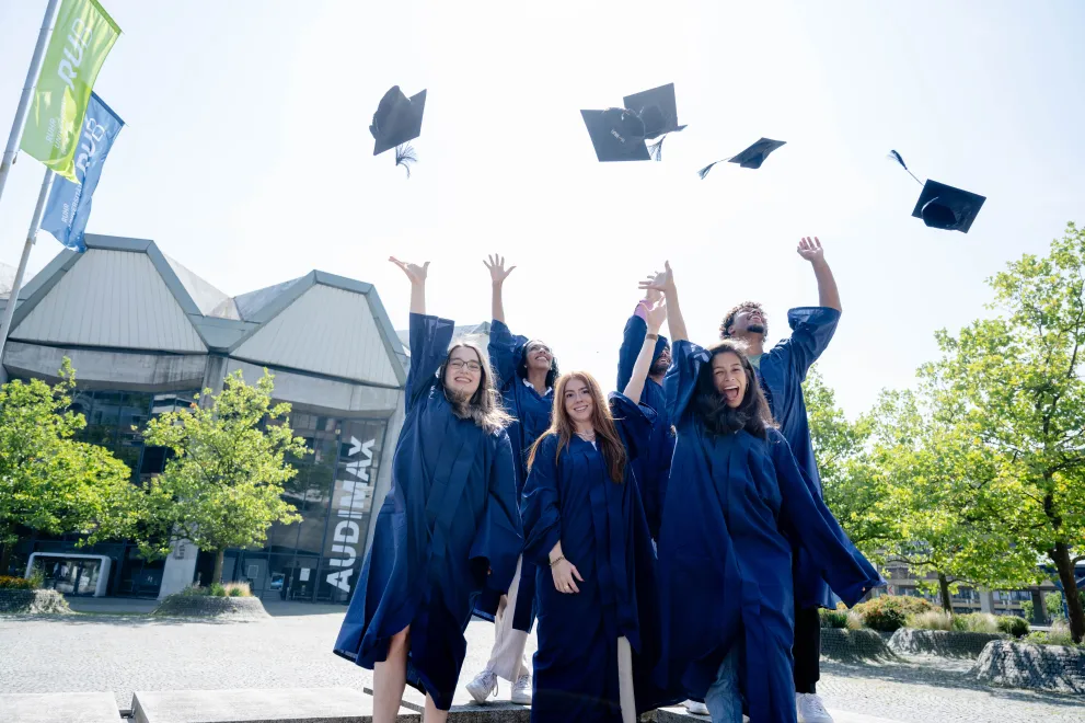 Foto: Mehrere Personen in blauen Talaren stehen auf dem Forums-Platz vor dem Audimax und werfen Hüte in die Luft. Die Hände sind in die Luftgestreckt, die Personen lachen und jubeln.