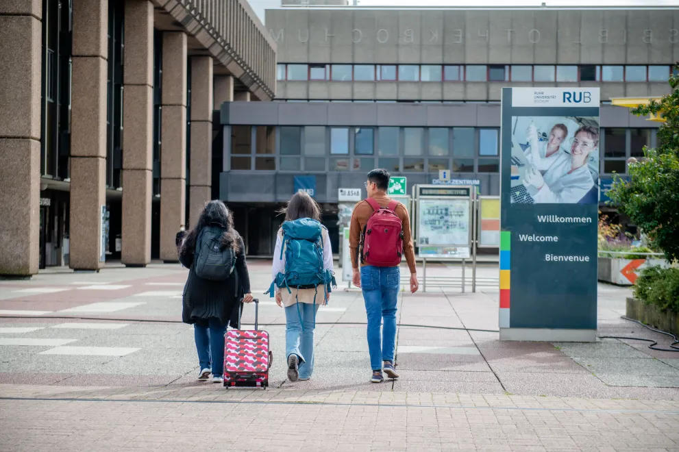 Drei Studierende betreten den Campus der RUB. Neben ihnen steht auf einem Schild "Wilkommen", "Welcome", und "Bienvenue".