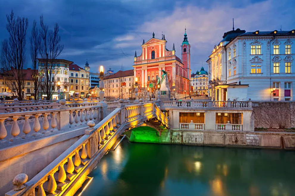 Eine gelb beleuchtete Brücke, die zu einer pinken Kirche in Ljubljana hinführt.